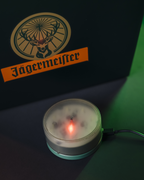 Jägermeister-logo op een donkere achtergrond met een klein rond apparaat dat licht uitstraalt.