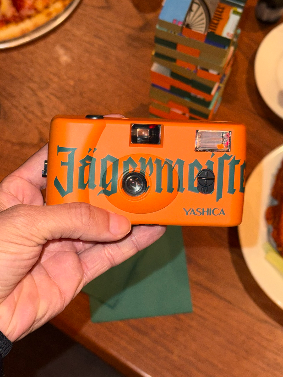 Oranje camera met 'Jägermeister'-branding vastgehouden door een hand op een houten tafel.