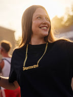 Vrouw in een zwart shirt en een gouden ketting met een Jägermeister-bedel, glimlachend buiten op een festival.