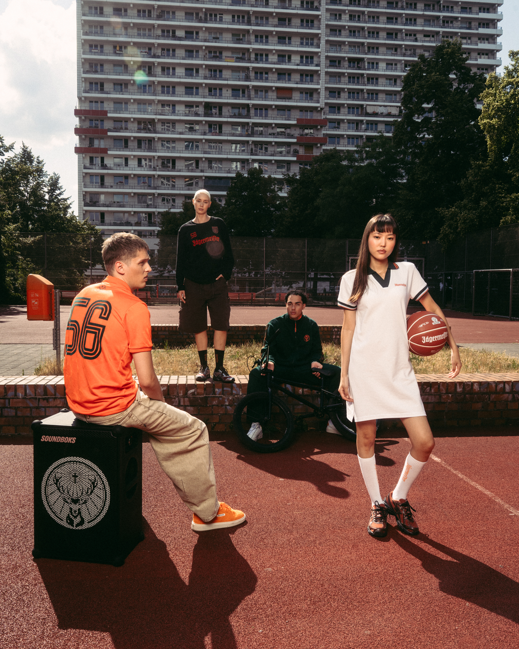 Group of people on an outdoor basketball court, featuring a person in a white Jägermeister polo dress holding a basketball, alongside others wearing Jägermeister apparel, with a high-rise building in the background.