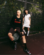 Person wearing a white Jägermeister polo dress standing beside a chain-link fence, with another person seated in front wearing a black Jägermeister sweater and shorts. Outdoor urban setting with greenery in the background.