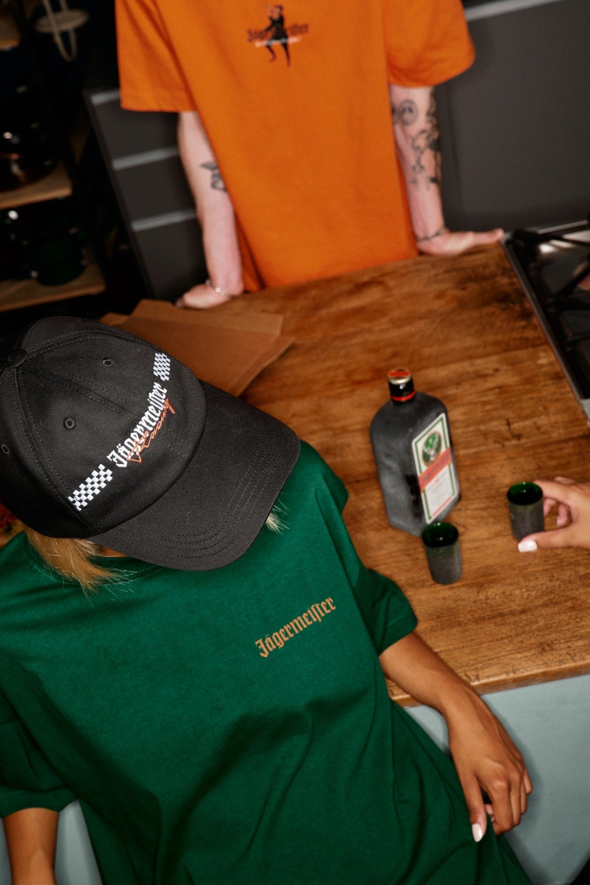 Persoon in een groen 'Jägermeister'-shirt en zwarte Jägermeister Racing Cap-pet zit aan een tafel met Jägermeister-producten erop.
