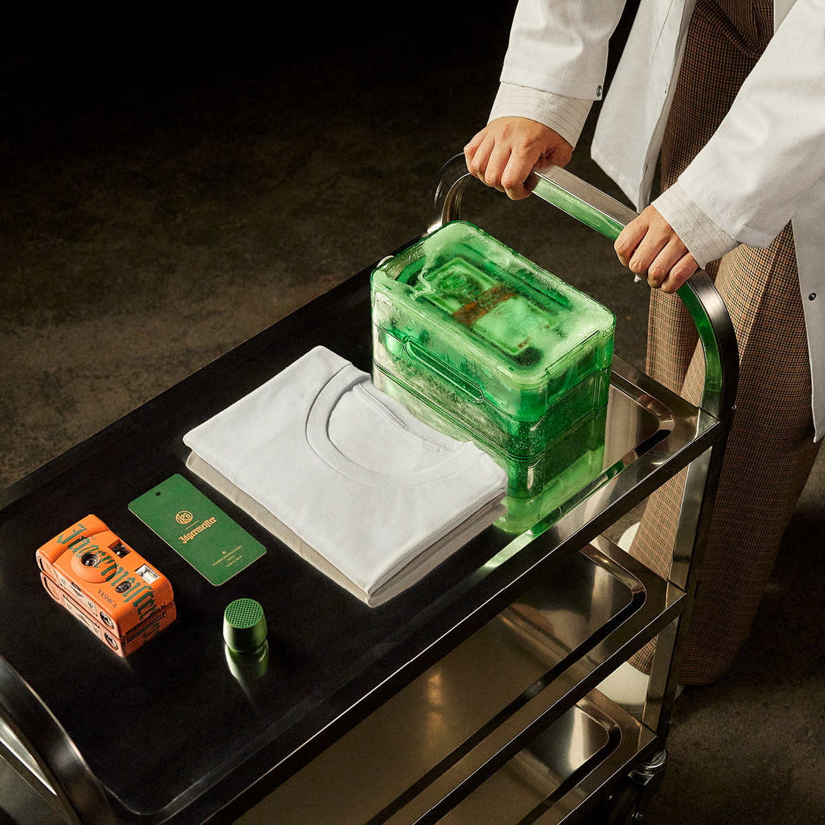 A person in a white lab coat pushes a black metal cart carrying a translucent green rectangular product box which includes a Jägermeister bottle, a folded white T-shirt, an orange analog camera and green fragrance paper, and a small green speaker in a dark studio setting.