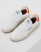 Een paar witte Jägermeister-sneakers met oranje accenten op de hiel en beige tussenzolen, gefotografeerd op een strakke witte studi achtergrond. De merknaam 'Jägermeister' is zichtbaar op de zijkant van de schoen.