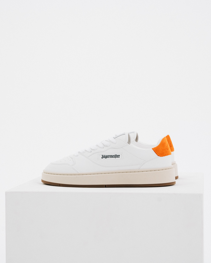 Een witte Jägermeister-sneaker met een feloranje hiellus en zwarte logotekst op de zijkant, tentoongesteld op een witte sokkel tegen een witte achtergrond.