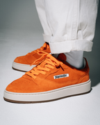 Een persoon die feloranje Jägermeister-sneakers draagt met een witte broek met omgeslagen pijpen, gefotografeerd vanaf de knieën omlaag op een lichtgrijze studi achtergrond. Op de schoen staat een wit zijlabel met 'Jägermeister' en een witte tussenzool met een gumkleurige buitenzool.