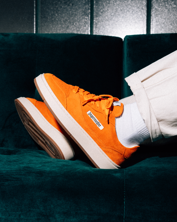 Feloranje Jägermeister sneakers met witte zolen en witte sokken, gedragen door een persoon die op een donkerblauwe fluwelen bank zit. Op het zijlabel van de schoen staat 'Jägermeister'.