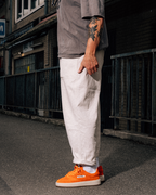 Een persoon staat buiten op straat en draagt een losse witte broek, een grijs T-shirt en feloranje lage sneakers. De foto is van borsthoogte naar beneden bijgesneden, met de rechterarm van de persoon zichtbaar en op de achtergrond een donkere stedelijke steeg of straatsetting.