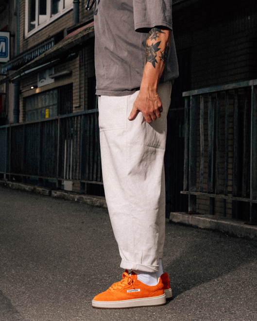Een persoon staat buiten op straat en draagt een losse witte broek, een grijs T-shirt en feloranje lage sneakers. De foto is van borsthoogte naar beneden bijgesneden, met de rechterarm van de persoon zichtbaar en op de achtergrond een donkere stedelijke steeg of straatsetting.
