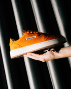 Een hand houdt een oranje Jägermeister sneaker vast met een witte zool en een zichtbaar 'Jägermeister'-label, tegen een donkere achtergrond met verticale metalen lamellen.