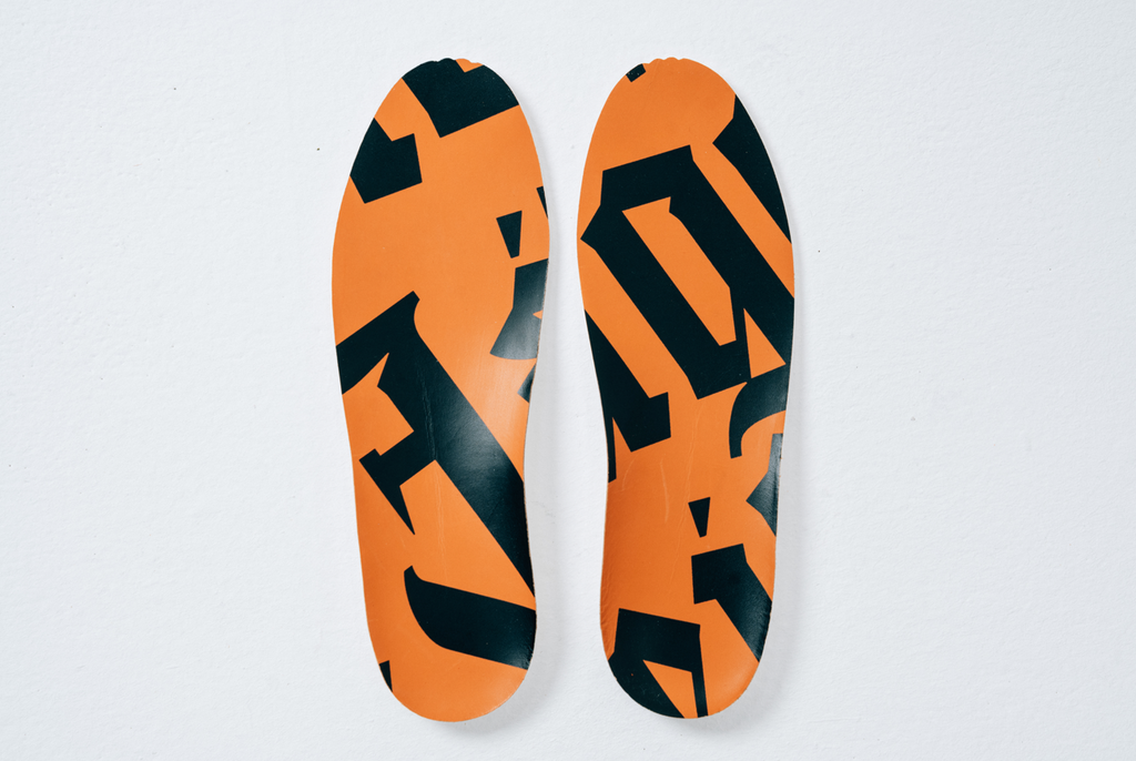 Een paar inlegzolen voor sportschoenen met een Jägermeister-oranje en zwart patroon, van bovenaf gezien op een lichte achtergrond.