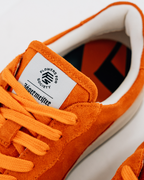 Close-up van een oranje Jägermeister sneaker met bijpassende oranje veters, een wit tonglabel met 'Flowers For Society' en 'Jägermeister', en een witte zijstreep op een witte achtergrond.