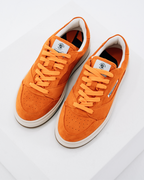 Een paar Jägermeister sneakers in levendig oranje, met bijpassende oranje veters, witte tussenzolen en witte binnenvoering, gefotografeerd op een strakke witte geometrische achtergrond.