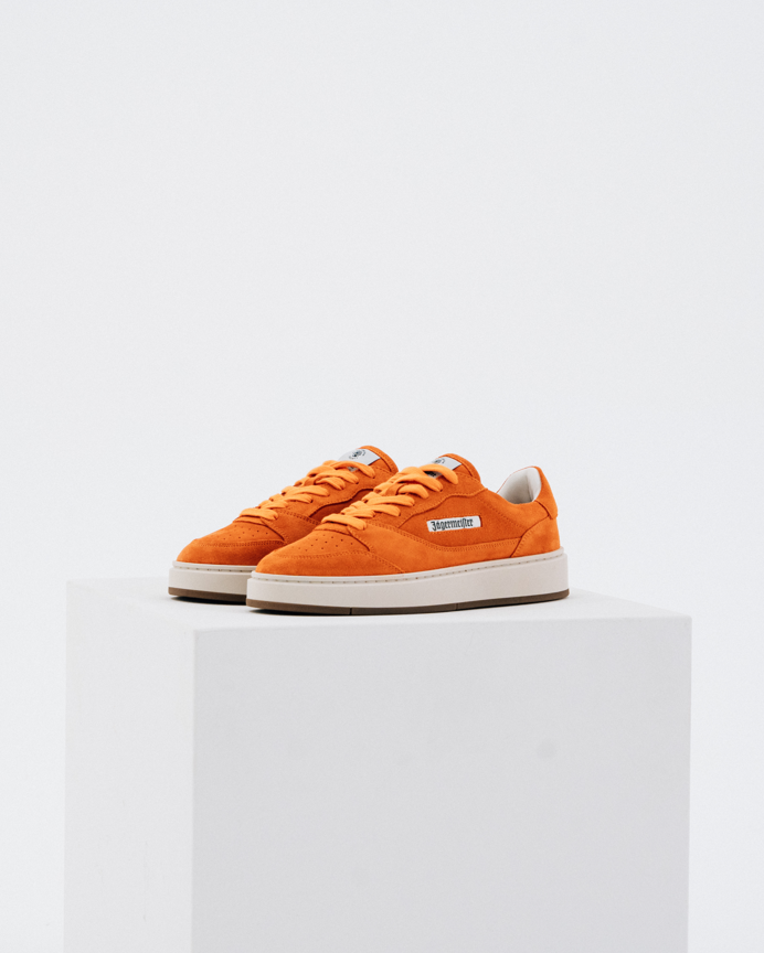 Een paar oranje Jägermeister sneakers met witte tussenzolen en gumzolen, weergegeven op een witte geometrische sokkel tegen een lichtgrijze achtergrond.