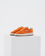 Een paar oranje Jägermeister sneakers met witte tussenzolen en gumzolen, weergegeven op een witte geometrische sokkel tegen een lichtgrijze achtergrond.
