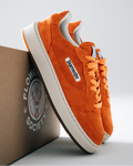 Oranje Jägermeister sneakers met witte zolen en een wit zijlabel met 'Jägermeister', weergegeven met een bruine doos bedrukt met 'Flower Society' op een lichtgrijze achtergrond.