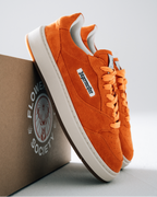 Oranje Jägermeister sneakers met witte zolen en een wit zijlabel met 'Jägermeister', weergegeven met een bruine doos bedrukt met 'Flower Society' op een lichtgrijze achtergrond.