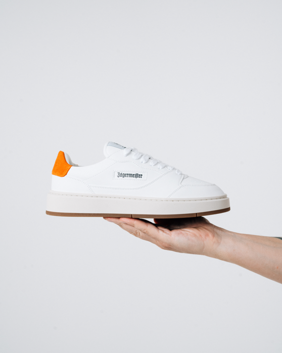 Een witte Jägermeister-sneaker met een feloranje hiellus en een zwart 'Jägermeister'-woordmerk op de zijkant, omhooggehouden door iemands hand tegen een lichte achtergrond.
