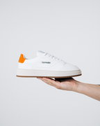 Een witte Jägermeister-sneaker met een feloranje hiellus en een zwart 'Jägermeister'-woordmerk op de zijkant, omhooggehouden door iemands hand tegen een lichte achtergrond.