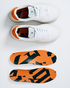 Een paar witte Jägermeister-sneakers van bovenaf, met daaronder twee bijpassende inlegzolen. De inlegzolen hebben een opvallend oranje en zwart abstract Jägermeister-woordmerkpatroon.