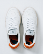 Een paar witte Jägermeister-sneakers gezien van bovenaf op een lichtgrijze achtergrond. De schoenen hebben witte veters, een wit leren bovenwerk en een feloranje hielaccent.