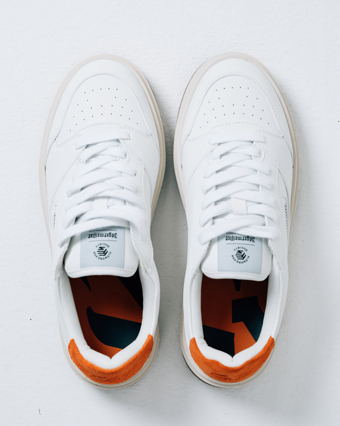 Een paar witte Jägermeister-sneakers gezien van bovenaf op een lichtgrijze achtergrond. De schoenen hebben witte veters, een wit leren bovenwerk en een feloranje hielaccent.