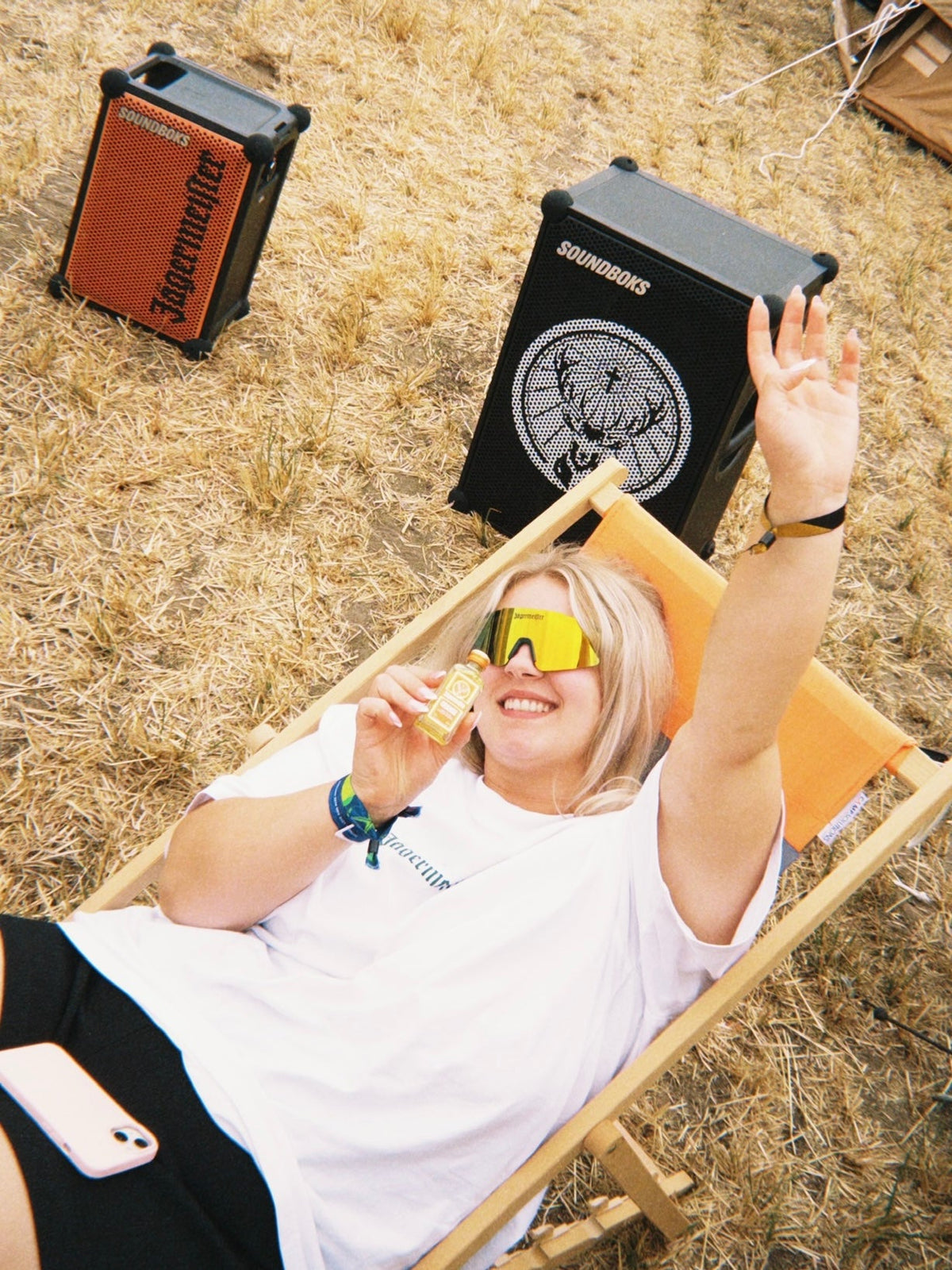 Een vrouw in een wit T-shirt en een zonnebril met geel getinte glazen ontspant buiten op een houten loungestoel op droog gras, met twee Soundboks-speakers zichtbaar achter haar.
