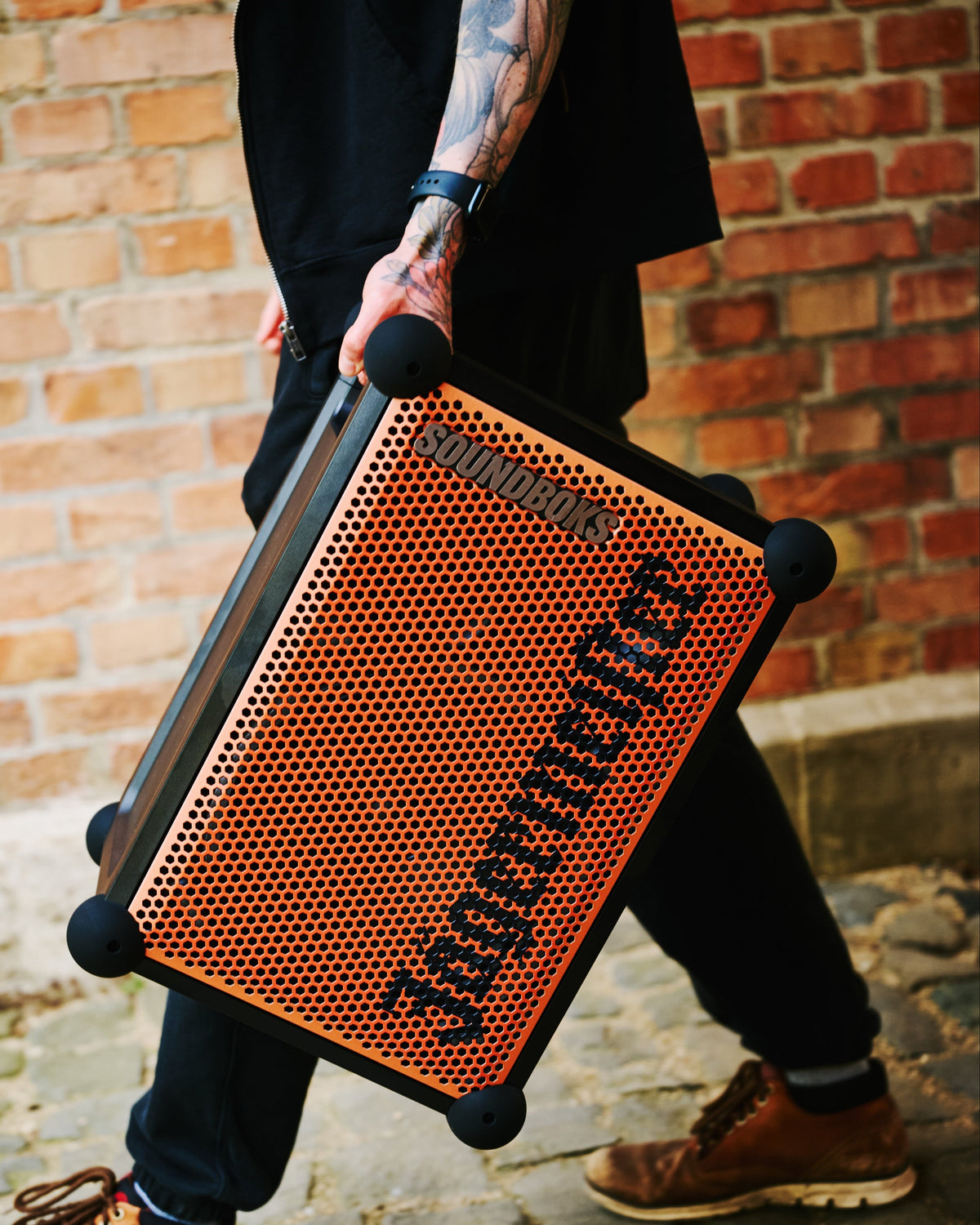 Person holding an orange and black portable Soundboks speaker with 'Jägermeister' branding in front of a brick wall.