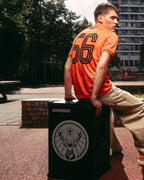 Een man in een oranje Jägermeister-jersey en beige broek zit op een zwarte Soundboks-speaker met Jägermeister-logo