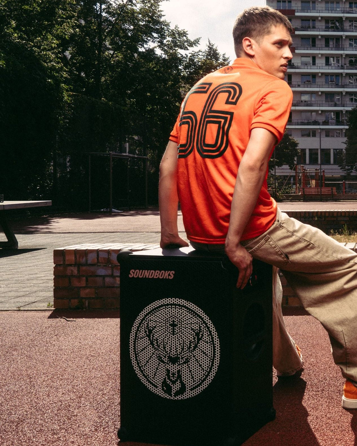 Een man in een oranje Jägermeister-jersey en beige broek zit op een zwarte Soundboks-speaker met Jägermeister-logo