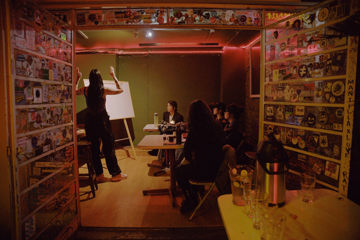 A dimly lit indoor meeting or workshop space with several people seated at tables, one person standing near a whiteboard, and walls covered in many small printed items. The scene is warm orange-red ambient lighting and appears to document an event or room rather than a clearly isolated sellable product.