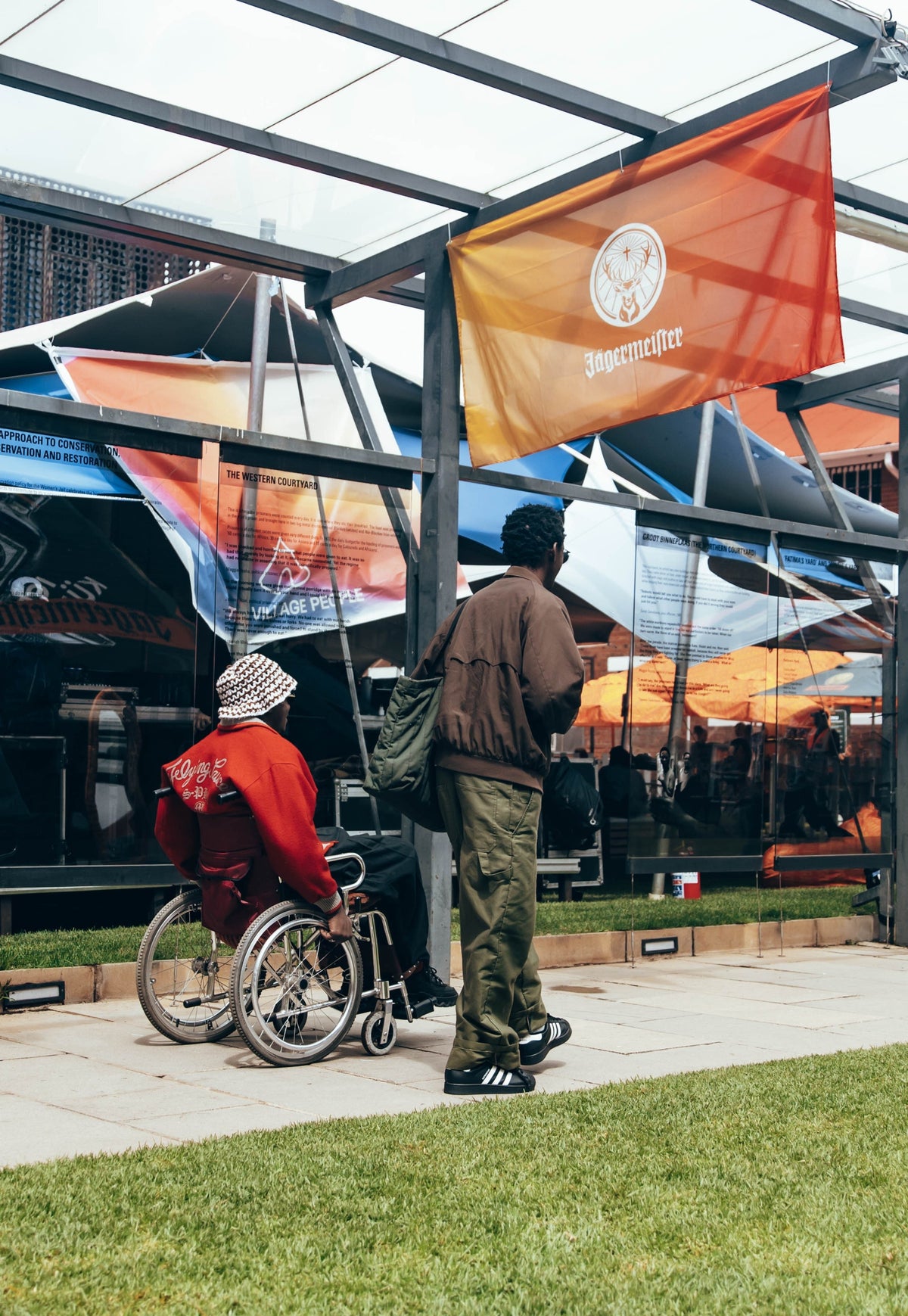 A person in a wheelchair wearing a red jacket and patterned headscarf sits beside another person in a brown jacket and green pants, standing outside a modern glass pavilion with an orange Jägermeister flag hanging above them.