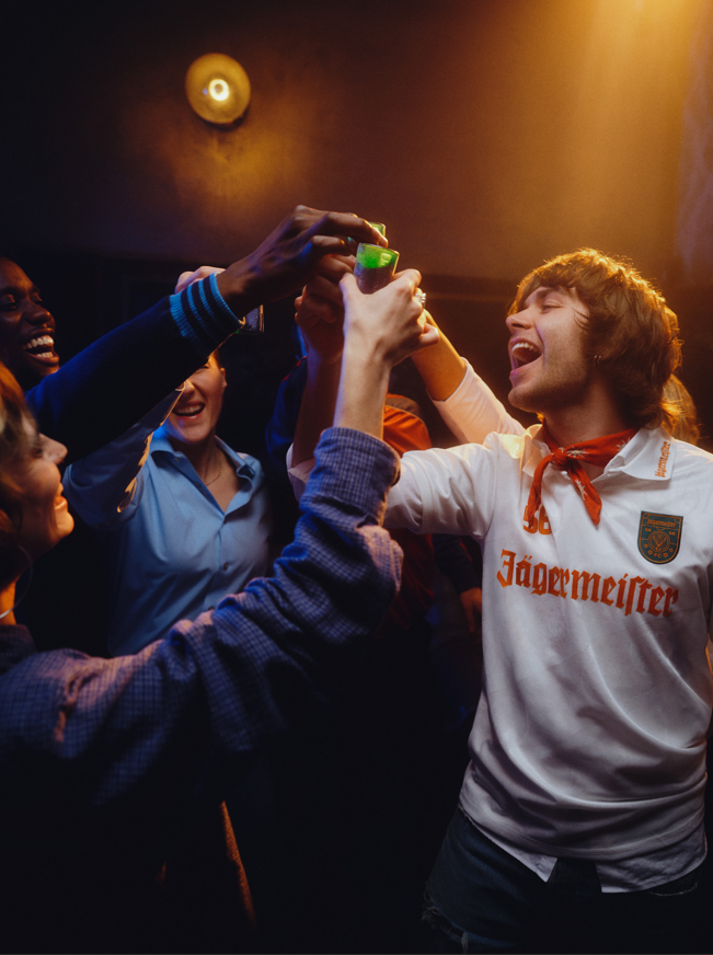 Een groep mensen viert feest met een persoon in een Jägermeister-shirt in een schemerig verlichte ruimte.